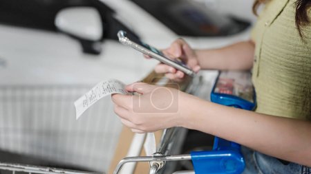 Frauen benutzen Handy, um Rechnung nach dem Einkauf im Supermarkt zu berechnen, Nahaufnahme der Hand überprüft lange Lebensmittelquittungen, sie verwendet eine Budgetierung und Geld sparende Handy-App