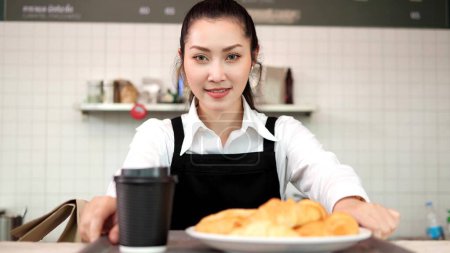 Foto de Hermosa mujer asiática barista está sosteniendo café caliente y pan sonriendo mira a la cámara mientras está de pie cerca del mostrador del bar en la cafetería - Imagen libre de derechos