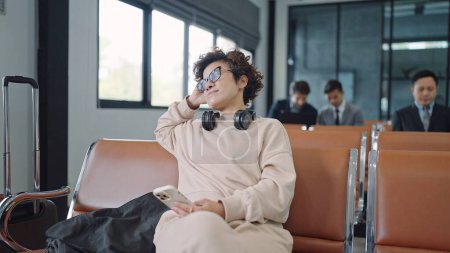 Foto de Turistas asiáticas sentadas y esperando el vuelo en la terminal del aeropuerto con smartphone y auriculares - Imagen libre de derechos