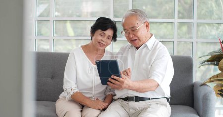 Foto de Feliz sonriente Asiática senior pareja de ancianos hablando en vídeo en la tableta mientras está sentado en el sofá en la habitación. Pareja de edad alegre saludando a la cámara mientras videochat en el dispositivo. Concepto casero - Imagen libre de derechos