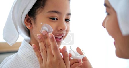 Téléchargez les photos : Mère asiatique ajoutant crème de traitement sur la joue à la petite fille avec robe de spa et la tête couverte d'une serviette blanche sur le lit ensemble à la maison - en image libre de droit
