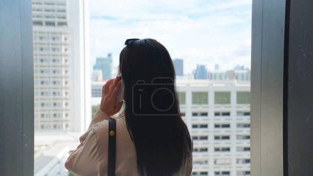 Photo for Back view of businesswoman in a suit talking on the phone Standing near a large glass window looking the city view - Royalty Free Image