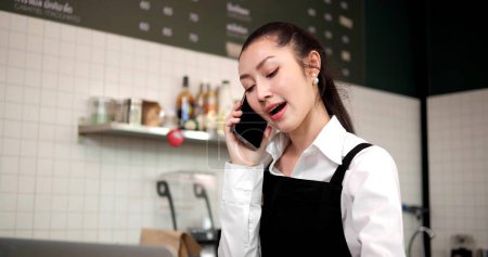 Photo for Asian woman coffee shop employee barista working at cafe. Smiling female waitress cashier taking order coffee and bakery from customer on phone. Small business owner and part time job working concept - Royalty Free Image