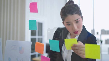 Foto de Empresaria asiática usando notas de papel pegajoso para compartir idea en la pared de vidrio en la oficina - Imagen libre de derechos