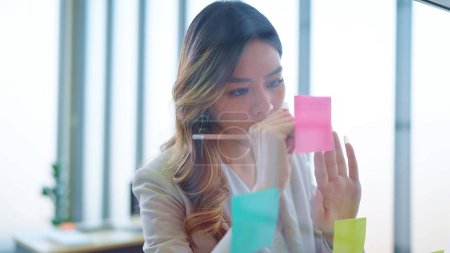 Téléchargez les photos : Femme d'affaires asiatique debout et écrire des tâches de projet sur le poste il notes collantes sur le mur de verre dans le bureau - en image libre de droit