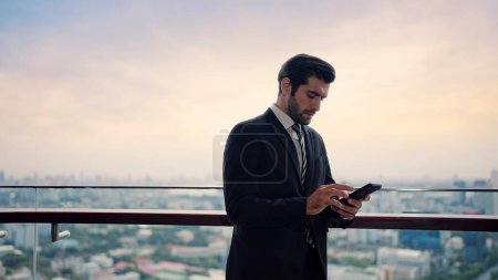 Foto de Hombre de negocios barbudo guapo escribiendo en el teléfono inteligente charlando con el cliente, de pie en la azotea desde el edificio de oficinas y vista de la ciudad - Imagen libre de derechos
