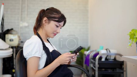 Photo for Asian young woman hairdresser with black apron using smartphone chatting with customer for booking at hair salon - Royalty Free Image