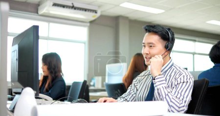 Foto de Hombre asiático es agente de atención al cliente o centro de llamadas con auriculares funciona en la computadora de escritorio mientras apoya al cliente en la llamada telefónica - Imagen libre de derechos
