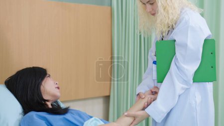 Téléchargez les photos : Femme blanche médecin prise de femme asiatique poignet patient pour mesurer le pouls couché sur le lit à la clinique ou à l'hôpital - en image libre de droit