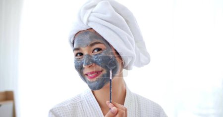 Foto de Mujer asiática aplicando máscara gris en la cara sonriendo a la cámara. Spa procedimientos de belleza en el hogar para chica. Concepto hidratante, hidratante y cuidado de la piel - Imagen libre de derechos