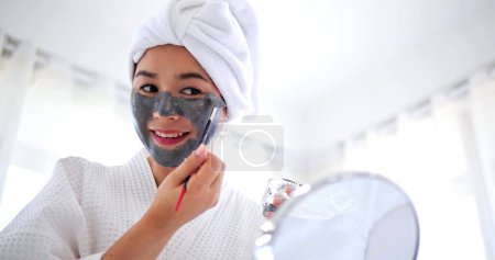 Téléchargez les photos : Femme asiatique appliquant un masque gris sur le visage souriant à la caméra. Spa procédures de beauté à la maison pour fille. Concept soin, hydratant et hydratant - en image libre de droit