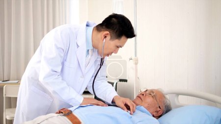 Foto de Médico masculino asiático que usa estetoscopio para escuchar el sonido pulmonar y cardíaco de un anciano asiático que está enfermo y acostado en la cama del hospital. concepto de apoyo sanitario a las personas mayores - Imagen libre de derechos