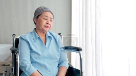 Téléchargez les photos : Patiente âgée asiatique assise en fauteuil roulant dans une salle de soins à domicile ou à l'hôpital devant la fenêtre avec rideau et pensée. Une vieille femme seule qui passe la journée seule à l'hôpital. Vieillesse, handicap - en image libre de droit
