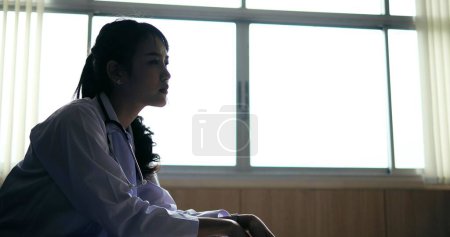 Photo for Young Asian depressed woman doctor sitting alone on chair stressed and anxious worried about patient treatment at hospital - Royalty Free Image