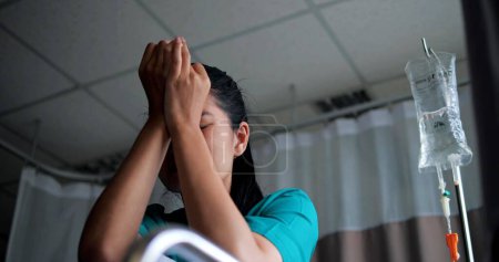 Téléchargez les photos : Jeune femme asiatique déprimée assise en fauteuil roulant ferme les yeux et sanglote à l'hôpital. Elle a souligné et anxieux inquiet sa santé - en image libre de droit