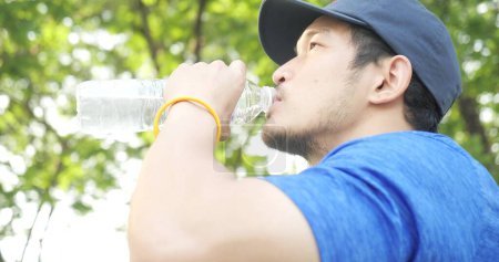 Foto de Guapo de hombre asiático beber agua de la botella después de hacer ejercicio en el parque verde con fondo de rayos de luz en el día soleado - Imagen libre de derechos