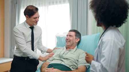 Téléchargez les photos : Un fils heureux rend visite à un vieux père en convalescence couché sur le lit à l'hôpital. et ils ont parlé avec le docteur - en image libre de droit