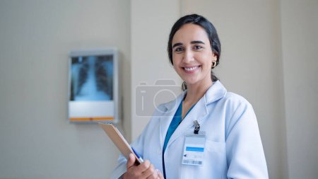 Photo for Smiling young hispanic latin woman doctor in white medical coat holding clipboard while standing and looking at camera in hospital - Royalty Free Image