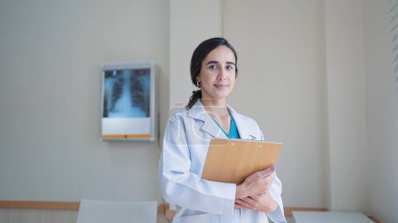 Photo for Young hispanic latin woman doctor in white medical coat holding clipboard while standing and looking at camera in hospital - Royalty Free Image