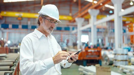 Foto de Ingeniero gerente líder hombre con casco escribiendo en el teléfono inteligente de pie en el área del lugar de trabajo en la fábrica de fabricación - Imagen libre de derechos