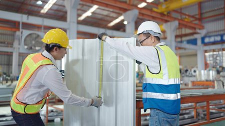 Téléchargez les photos : Deux travailleurs asiatiques en uniforme de sécurité travaillant à la fabrication dans une usine métallique et mesurant la taille du toit métallique - en image libre de droit