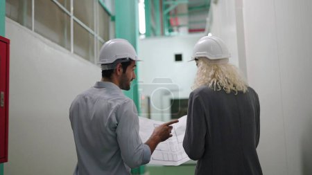 Photo for Back view of two industry engineers looking project blueprints discussing while walking in large distribution warehouse center - Royalty Free Image