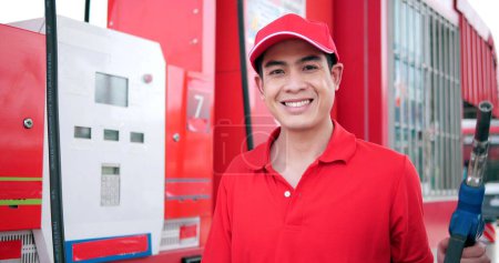 Foto de Retrato del trabajador asiático en uniforme rojo con la boquilla de la bomba de combustible que mira la cámara en el servicio de gasolinera - Imagen libre de derechos