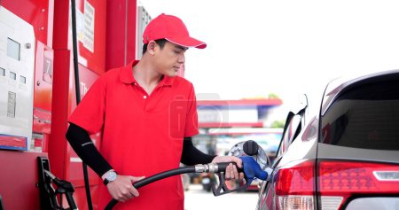 Foto de Trabajadora asiática empleada en ropa de trabajo repostaje uniforme para automóvil con boquilla de llenado de gasolina en la bomba de la gasolinera - Imagen libre de derechos