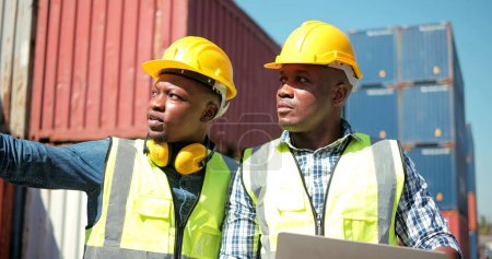 Téléchargez les photos : Deux ingénieurs africains professionnels portant un uniforme de sécurité et des chapeaux durs travaillant sur un ordinateur portable et tenant une radio de communication au cargo - en image libre de droit