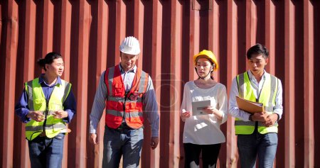 Téléchargez les photos : Groupe de travailleurs ingénieurs équipe portant le casque dur de sécurité parler et marcher à fond de la cour de conteneurs de fret logistique - en image libre de droit