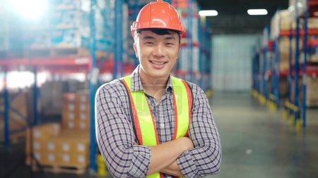 Téléchargez les photos : Travailleur asiatique debout dans un grand centre de distribution d'entrepôt avec les bras croisés dans des étagères avec fond de marchandises. Travailleur sentant et regardant vers la caméra. Industrie logistique - en image libre de droit