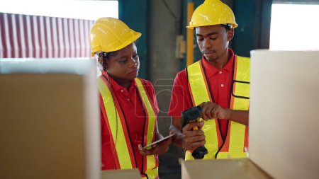 Foto de Dos trabajadores jóvenes afroamericanos revisando el estado del stock y escaneando el código de barras en la caja de cartón en el almacén de la industria de la fábrica de negocios usando traje de ingeniero y casco para mayor seguridad - Imagen libre de derechos