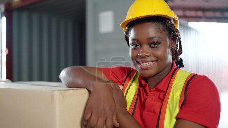 Téléchargez les photos : Jeune travailleuse noire afro-américaine regardant la caméra portant une combinaison d'ingénieur et un casque pour la sécurité. en entrepôt, concept d'entreprise d'exportation internationale - en image libre de droit