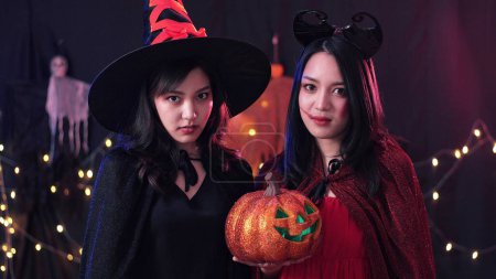 Photo for Two Asian woman in Halloween concept costume and looking at camera on over dark backdrop with light bokeh background - Royalty Free Image