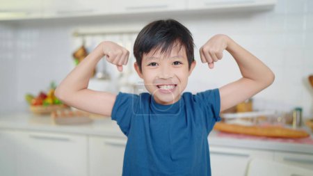 Photo for Asian little cute boy in blue shirt demonstrating his muscles or biceps at kitchen room. Boy power future and dream concept - Royalty Free Image