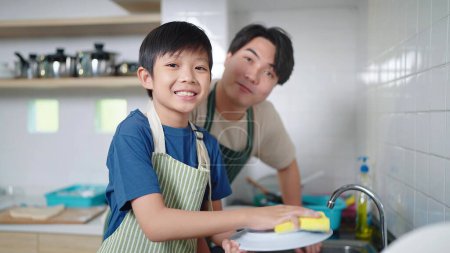 Foto de Feliz asiático familia, pequeño hijo y padre son lavado platos en la cocina y mirando a cámara - Imagen libre de derechos