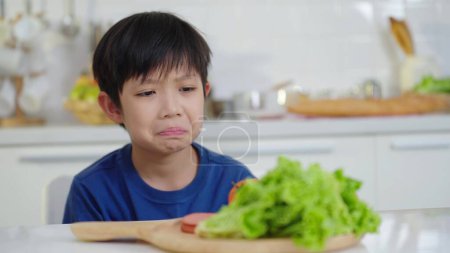 Téléchargez les photos : Les enfants n'aiment pas manger de légumes. Le visage malheureux du garçon ne veut pas manger des légumes sains - en image libre de droit
