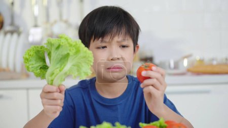 Téléchargez les photos : Visage malheureux de petit garçon asiatique tenant tomate et légumes verts refusant de manger regardant avec dégoût - en image libre de droit