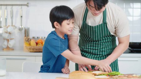 Foto de Asiático hijo ayudando a padre preparando sándwich para el desayuno en vacaciones en la sala de cocina - Imagen libre de derechos