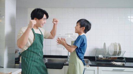 Téléchargez les photos : Père asiatique et petit fils dans tablier regardant heureux après avoir lavé la vaisselle dans la salle de cuisine. Concept de style de vie familial - en image libre de droit