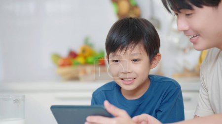 Téléchargez les photos : Père asiatique enseignant petit fils en utilisant une tablette numérique pour apprendre en ligne à la maison dans la distance sociale pendant l'auto quarantaine. Concept d'éducation en ligne - en image libre de droit
