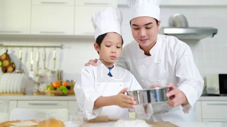 Photo for Asian father teaching little son preparing dough, baking cookies in kitchen wearing chef costume enjoying weekend playtime cooking baking. Happy family concept - Royalty Free Image