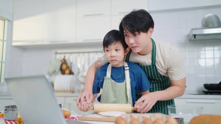 Foto de Asiático padre soltero enseñando poco hijo haciendo masa para el pan, aprendiendo de videos en las redes sociales utilizando labtop en la cocina - Imagen libre de derechos