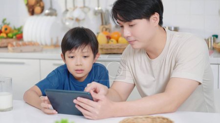 Téléchargez les photos : Asiatique père célibataire et petit fils utilisant tablette pour apprendre à cuisiner dans la salle de cuisine en vacances. Joyeux concept de famille. Concept de père unique - en image libre de droit