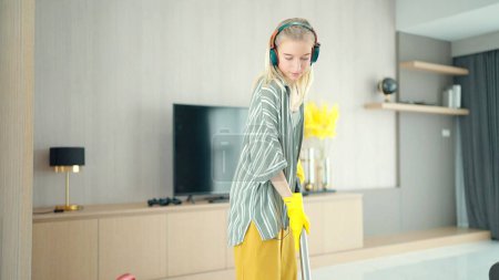 Foto de Mujer joven rubia limpiando piso con aspiradora moderna y bailando disfrutando de la música a través de auriculares en casa - Imagen libre de derechos