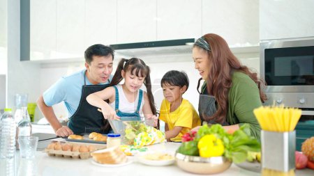 Photo for Happy Asian family in the kitchen cooking together healthy. Parents teach little children healthy habits and how to mix vegetables in salad bowl - Royalty Free Image