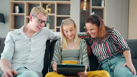 Téléchargez les photos : Heureux parents caucasiens et la fille adolescente s'amusent à utiliser un ordinateur portable à la maison ensemble, regarder un film ou naviguer sur Internet, assis sur le canapé dans le salon - en image libre de droit