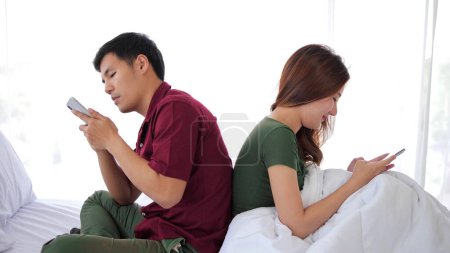 Foto de Un par de asiáticos sentados en la cama, enviando mensajes de texto en el teléfono móvil, se dieron la espalda el uno al otro, usando un teléfono inteligente. Indiferente y no prestan atención el uno al otro - Imagen libre de derechos