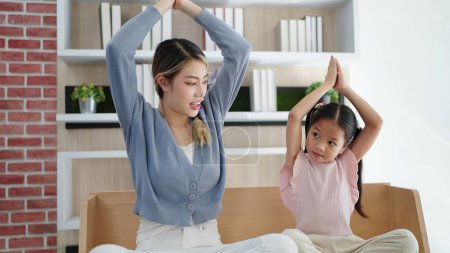 Foto de Feliz asiática mamá e hija haciendo yoga juntos en casa manteniendo colgando padmasana, equilibrio de brazos. La familia se sienta en posición de loto en el sofá en la sala de estar. Yoga en familia - Imagen libre de derechos