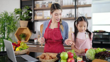 Photo for Happy family Asian mother and daughter cooking together with table full of fruits and vegetables in kitchen. Learning online cooking using laptop - Royalty Free Image